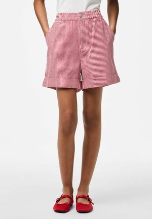 Person som har på seg røde og hvite shorts med vertikale striper og oppbrettede kanter, samt røde Mary Jane-sko, står med hendene i lommene.