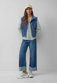 Gilet en denim avec deux poches avant, associé à une chemise à rayures vertes, un pantalon en denim à jambes larges et des baskets légères. Casquette neutre en accent.