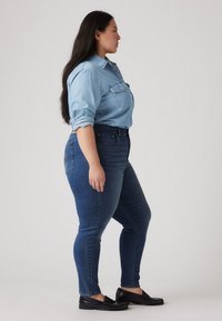 Camisa de ganga azul clara com colarinho abotoado, usada com jeans escuros de corte justo e mocassins pretos, vista de lado.