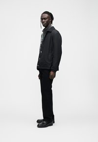 Svart jakke med knappeåpning og kontrasterende mønster på siden; kombinert med svarte bukser og svarte sko. Glatt stofftekstur.