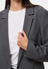 Blazer gris avec des revers structurés et une coupe décontractée, porté sur un t-shirt blanc. Tissu lisse avec peu de détails et lignes épurées.