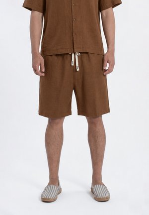 Uomo che indossa una camicia marrone con bottoni e pantaloncini abbinati con coulisse bianca, abbinati a scarpe slip-on a righe grigie e bianche.