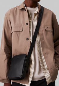 Černá kožená kabelka přes rameno se hladkým povrchem, obdélníkovým tvarem, minimálním logem a nastavitelného popruhem, nošena ve stylu crossbody.
