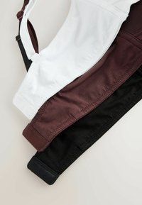 Trois soutiens-gorge en tissu doux de couleur blanche, marron et noire, empilés et posés à plat, montrant les bretelles réglables et les bords cousus.