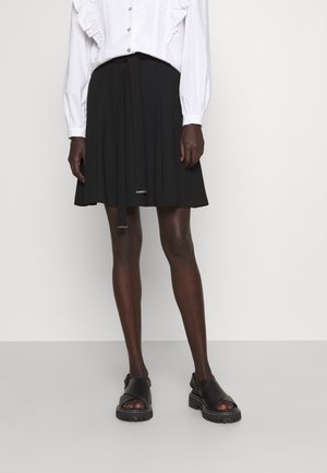 Osoba ubrana w czarną plisowaną spódnicę z przewiązanym paskiem, białą bluzkę z falbankami oraz czarne masywne sandały z odkrytymi palcami, stojąca na tle jednolitego tła.