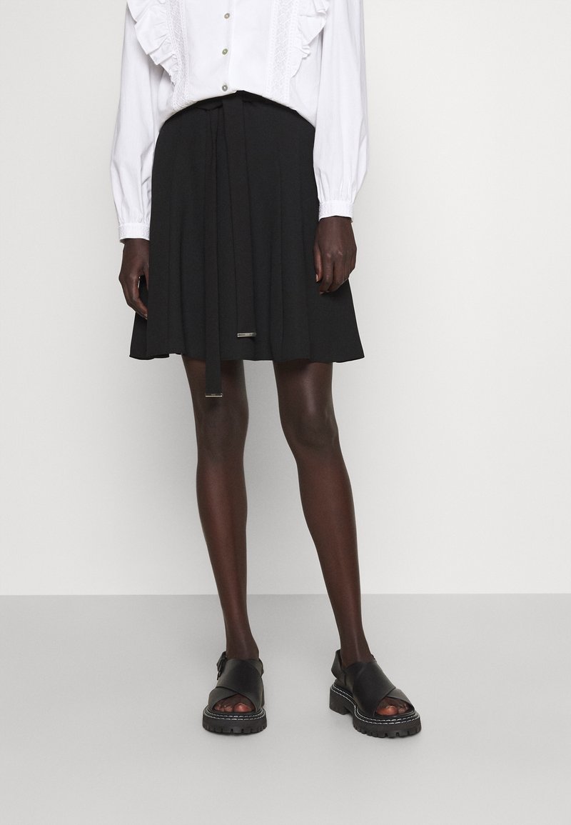 Personne portant une jupe plissée noire avec une ceinture nouée, un chemisier blanc à volants, et des sandales noires épaisses à bout ouvert, debout devant un fond uni.