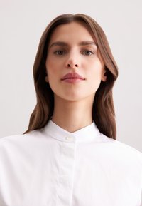 Camicia bianca con colletto, con chiusura a bottoni, realizzata in un tessuto liscio e testurizzato, dal design minimalista, con scollatura dritta e vestibilità su misura.