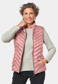 Gevoerde vest in lichtroze met een chevron-quiltpatroon, voorzien van een staande kraag en twee zijzakken. Draag het over een grijze gebreide trui.