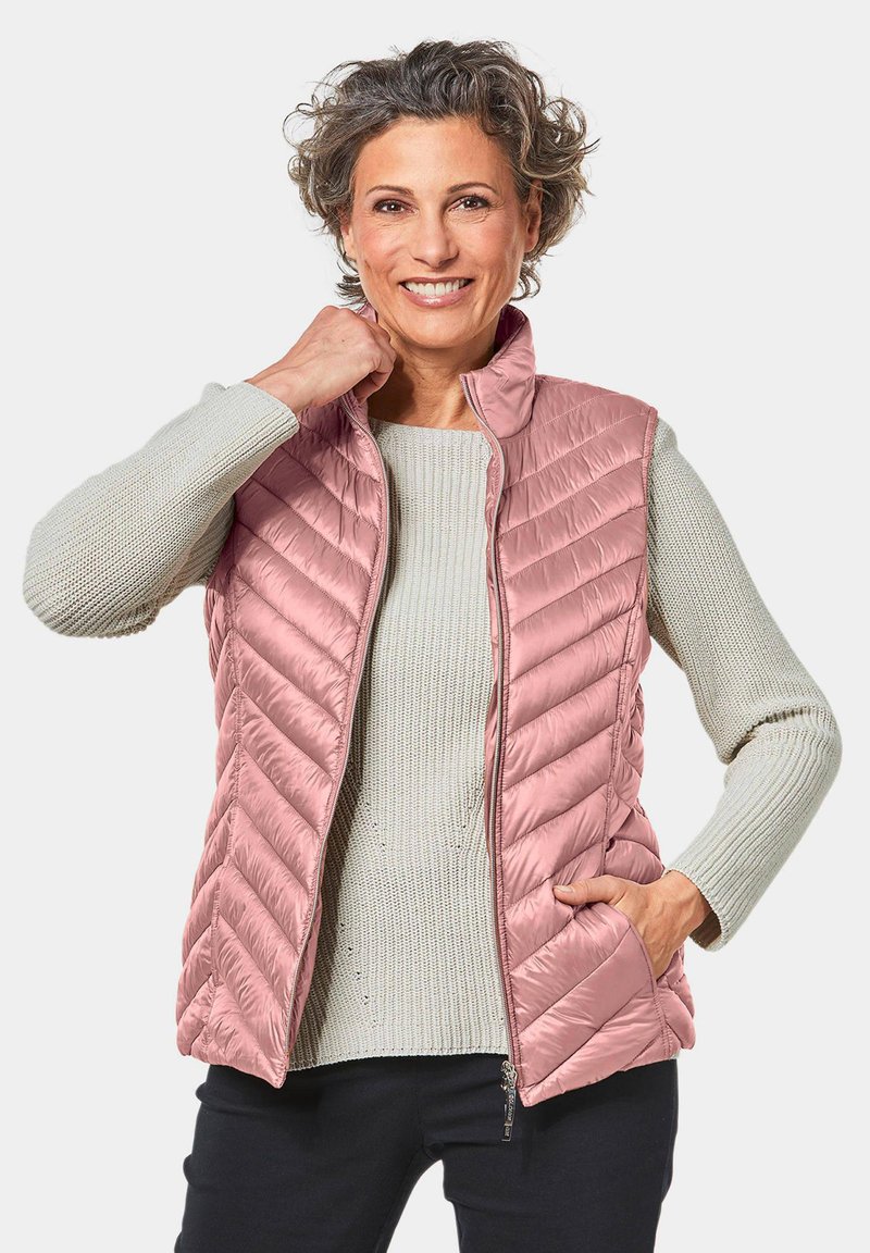 Gevoerde vest in lichtroze met een chevron-quiltpatroon, voorzien van een staande kraag en twee zijzakken. Draag het over een grijze gebreide trui.