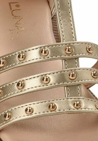 Sandalia de imitación cuero dorado con múltiples tiras adornadas con tachuelas metálicas, textura suave y plantilla acolchada.