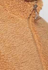 Fleecejacka i varm tan med en strukturerad yta. Har en dragkedja och en liten silverfärgad Nike-logocharm fäst vid dragkedjan.