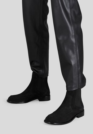 Stivaletti alla caviglia in suede nero con pannelli elastici laterali e un design elegante. La suola è nera lucida, con una silhouette minimalista e moderna.