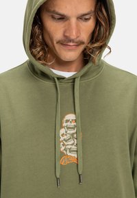 Felpa verde oliva con un grafico di teschi sovrapposti con accenti arancioni. I materiali sembrano morbidi, con cappuccio regolabile e polsini a coste.