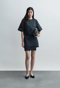 Melna divdaļīga apģērba komplekts, kas sastāv no kastveida īsām piedurknēm topa un pieguļoša svārka. Audums izskatās gluds ar minimālām šuvēm. Komplektā ar teksturētu rokassomu.