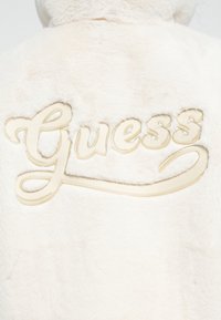 Vista posteriore di un giubbotto bianco e fluffy con il logo "Guess" ricamato beige sulla parte superiore della schiena.