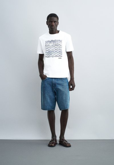 Άνδρας που φοράει λευκό κοντομάνικο πουκάμισο με κυματοειδές σχέδιο και μπλε τζιν σορτς, στέκεται με το ένα χέρι στην τσέπη και καφέ σανδάλια.