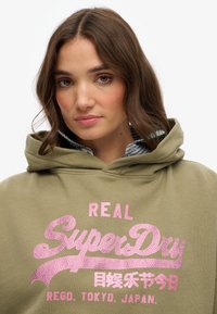 Felpa verde oliva in cotone, con un grande logo rosa glitterato "SuperDry". Include un cappuccio e una maglietta a righe blu a strati sotto.