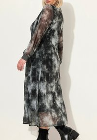 Długa czarna i szara sukienka tie-dye z przezroczystymi długimi rękawami, zwiewnym krojem i teksturowanymi mankietami. Noszona z czarnymi botkami za kostkę.