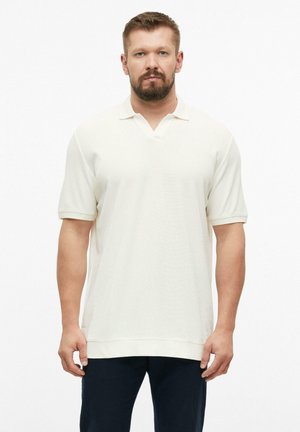 Mann mit kurzen braunen Haaren und Bart, der ein weißes kurzärmeliges Poloshirt und eine dunkle Hose trägt, steht vor einem schlichten weißen Hintergrund.