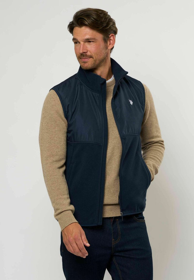 Marineblauw vest met een gladde, glanzende voorkant en fleece-materiaal. Heeft een hoge kraag, een rits aan de voorkant, zijzakken en een klein logo op de borst.