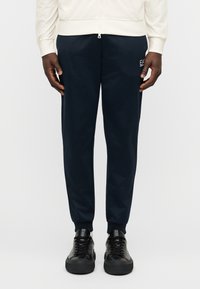 Παντελόνι jogger σε ναυτικό μπλε από απαλό ύφασμα, με ελαστικές μανσέτες, πλαϊνές τσέπες και μικρό λογότυπο της μάρκας στο μηρό. Συνδυασμένο με μαύρα αθλητικά παπούτσια.