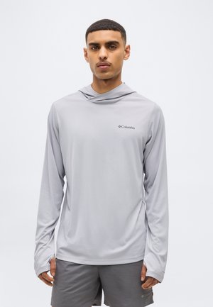 Noor mees, kes kannab helehalli Columbia pikkade varrukatega kapuutsiga särki pöidlahõlmadega ja tumedat halli lühikesi pükse, seisab valge tausta ees.