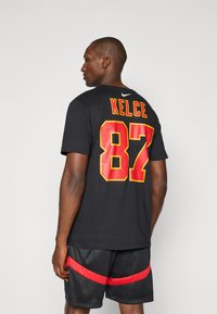 Sort kortærmet t-shirt med "KELCE" og "87" i fed rød og gul skrift på ryggen. Parret med sorte shorts med røde striber.