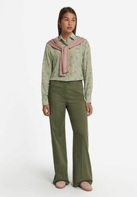 Lysegrøn bluse med blomster og knaplukning foran, sammen med olivengrønne bukser med bred ben. Lyserød sweater draperet om skuldrene og lyserøde sko.
