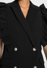 Svart dubbelknäppt blazer med volangärmar och dekorativa kluster av strasstenar, bärs av en person.