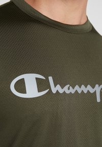 Olivově zelené sportovní tričko s texturedním vzorem a velkým bílým logem "Champion" umístěným na hrudi. Krátké rukávy a kulatý výstřih.