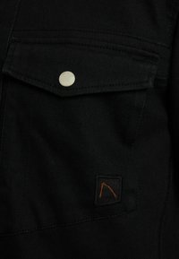 Poche en tissu noir avec rabat sécurisé par un bouton argenté portant l'étiquette "CHASIN,", présentant un petit patch noir avec un logo stylisé orange.