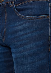 Zbliżenie na kieszeń ciemnoniebieskich dżinsów z pomarańczowym przeszyciem i metalowymi nitami.