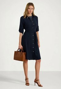 Γυναίκα σε ναυτική φόρεμα με κουμπιά και γυρισμένα μανίκια, κρατώντας καφέ δερμάτινη τσάντα, φορώντας ριγέ μπλούζα από κάτω και καφέ ψηλοτάκουνες σάντες.