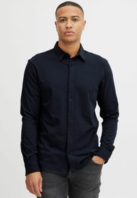 Hombre con una camisa de botones negra de manga larga y cuello, de pie contra un fondo blanco liso, con una mano en el bolsillo.
