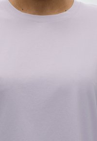 Κοντινό πλάνο ενός ατόμου που φοράει απλό, ανοιχτό μωβ μπλουζάκι με στρογγυλή λαιμόκοψη, εμφανίζοντας μόνο το λαιμό και το πάνω μέρος του στήθους.