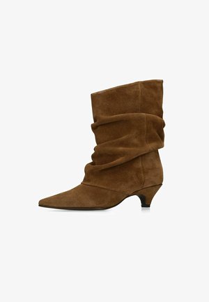 Bruine suède enkellaars met een slobberige bovenkant, spits toelopende neus en lage kittenhak op een witte achtergrond.