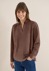 Femme aux cheveux bruns et raides souriant, portant une chemise marron à manches longues et un jean bleu, debout devant un fond beige.