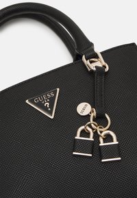 Détail d'un sac à main noir texturé avec doubles poignées, ferrures dorées, deux petits charms cadenas et une plaque triangulaire avec le logo Guess USA.