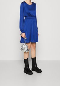 Robe en satin bleu à manches longues, taille smockée, associée à un sac à cordon argenté et des bottes à semelles épaisses noires. Fond uni.