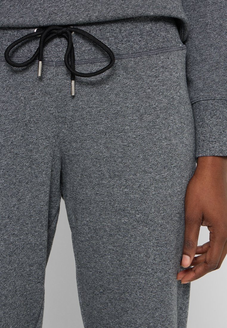 Zbliżenie na szare dresy z czarnym sznurkiem związanym w kokardkę, noszone przez osobę o ciemnej karnacji, z ręką luźno opadającą na bok.