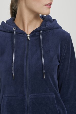 Mornarsko plava dukserica s patentnim zatvaračem, izrađena od mekane tkanine s prednjim džepom, ukrašena je bijelim detaljima na vezicama i metalnim patentnim zatvaračem.