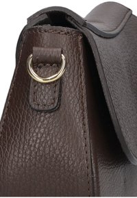 Hoek van een bruine leren handtas met een gestructureerd oppervlak, voorzien van een gouden metalen ring en een klep met zichtbare stiksels.