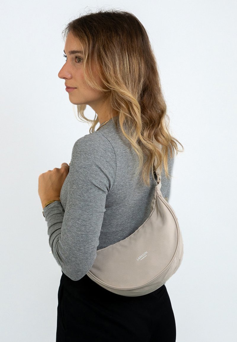 Donna con capelli ondulati alla spalla, che indossa un maglione grigio a maniche lunghe e pantaloni neri, mentre porta una borsa a tracolla a forma di mezzaluna beige.