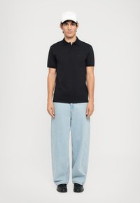 Svart pikétröja, kort ärm, slim fit. Ljusblå vida jeans, strukturerad tyg. Svarta skor. Vit basebollkeps med ventilationsdetaljer.