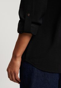 Czarna koszula z podwiniętymi mankietami z detalem guzika, w połączeniu z ciemnoniebieskimi dżinsami; gładka faktura i elegancki design.