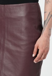 Burgunderfarget skinnskjørt med en glatt tekstur og rett design. En side søm er synlig, med en hånd som hviler på stoffet.