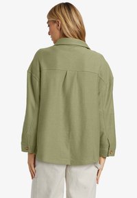 Olivgrön oversize-jacka med ett strukturerat tyg, kragdesigne och knappade manschetter, med en veck bak för extra detalj.