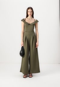 Olivengrønn jumpsuit med tettsittende bodice, av-skulderen design, og vide ben, laget av glatt stoff; kombinert med en svart veske.
