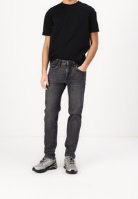 Černé tričko s krátkým rukávem zkombinované s úzkými tmavě šedými džíny. Model má na sobě šedé sportovní boty s černými akcenty.