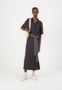 Robe chemise gris foncé à manches courtes, boutonnée à l'avant, cintrée avec une ceinture en chaîne, associée à des baskets blanches et un petit sac à bandoulière beige.